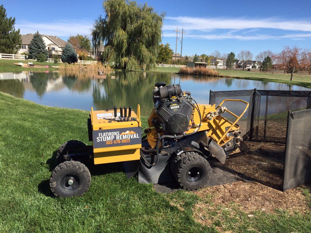 Yellow Flatirons Stump Removal grinder on a ground cloth next to steel mesh safety screens, beside a pond in Boulder